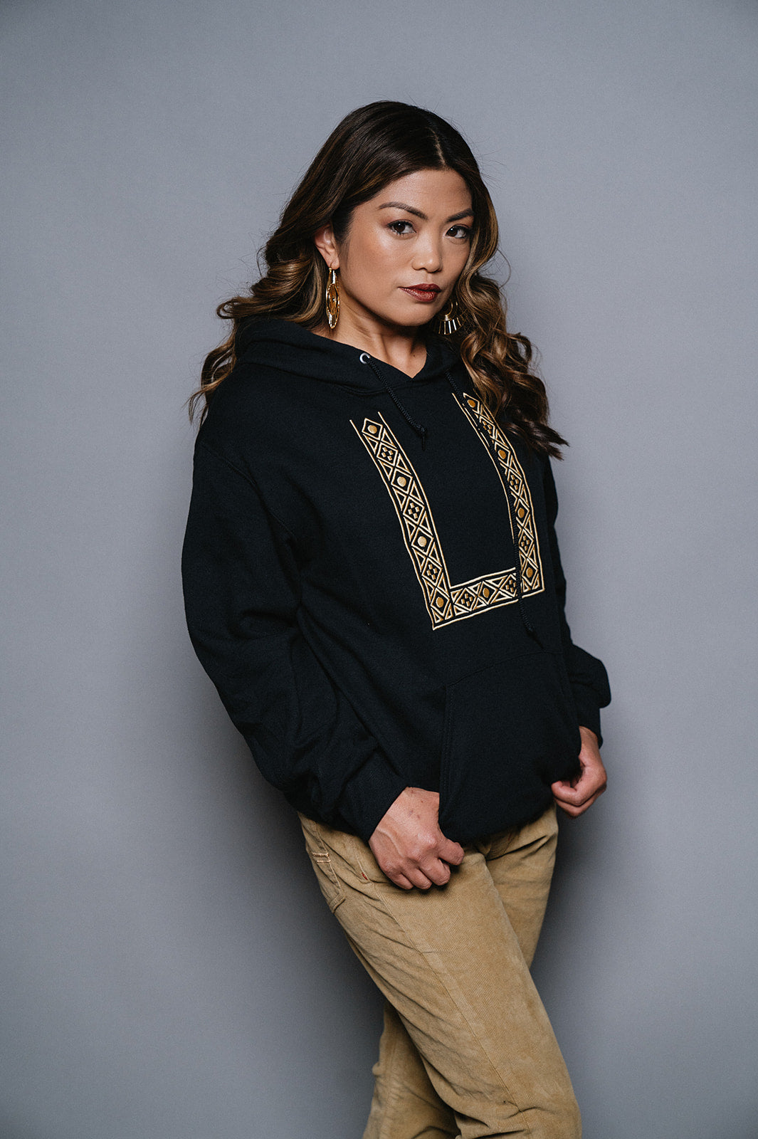 Filipina woman with dark brown hair with curls is wearing a black pullover, long sleeve hoodie with gold geometric embroidery resembling a barong but streetwear. She wears khaki colored corduray pants.