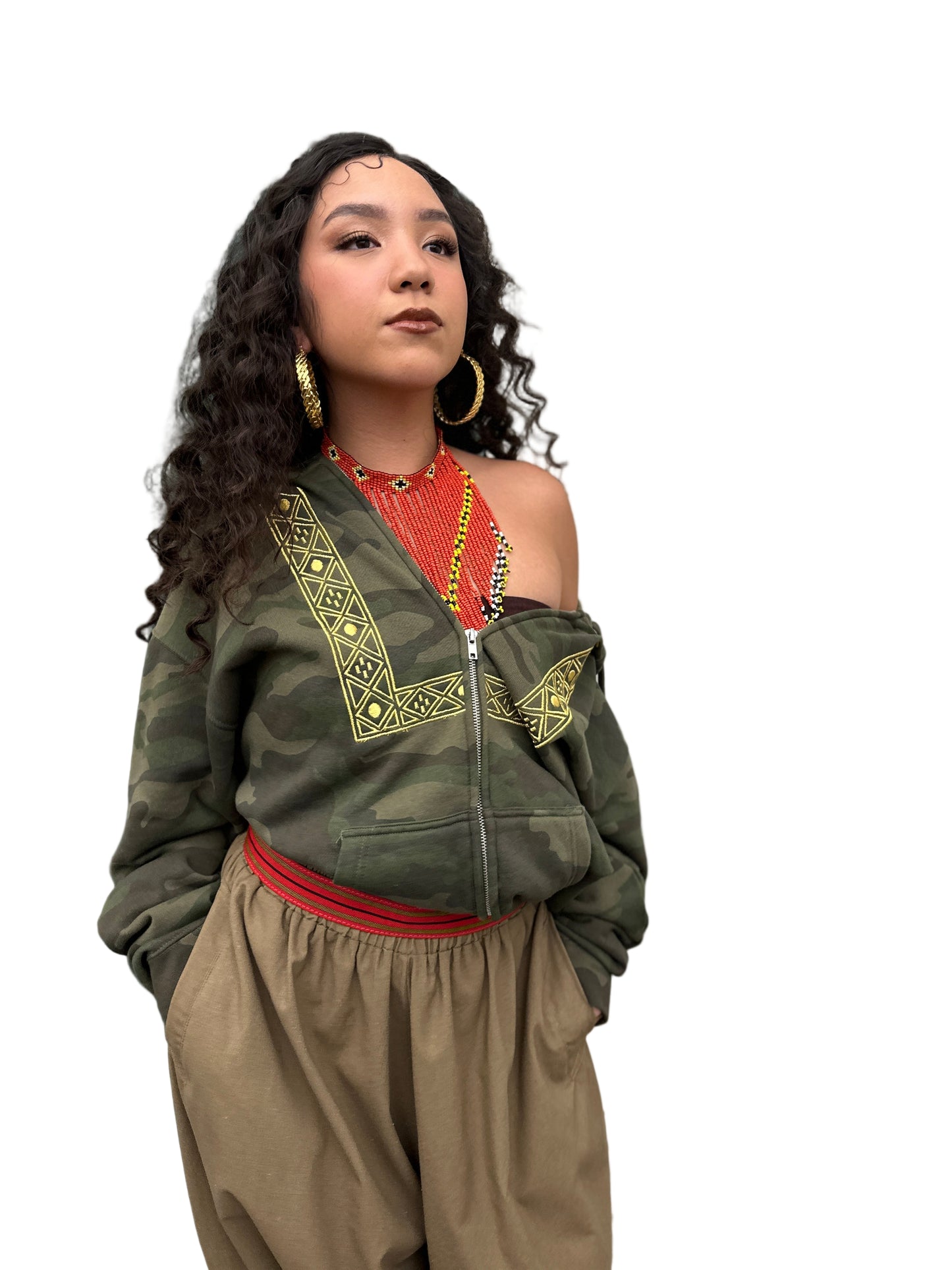 Woman wearing a camouflage zip up hoodie with gold embroidery, one side off the shoulder, with red and yellow beaded necklace, gold hoops and beige pants.