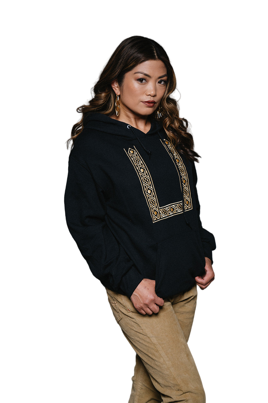 Filipina woman with dark brown hair with curls is wearing a black pullover, long sleeve hoodie with gold geometric embroidery resembling a barong but streetwear. She wears khaki colored corduray pants.