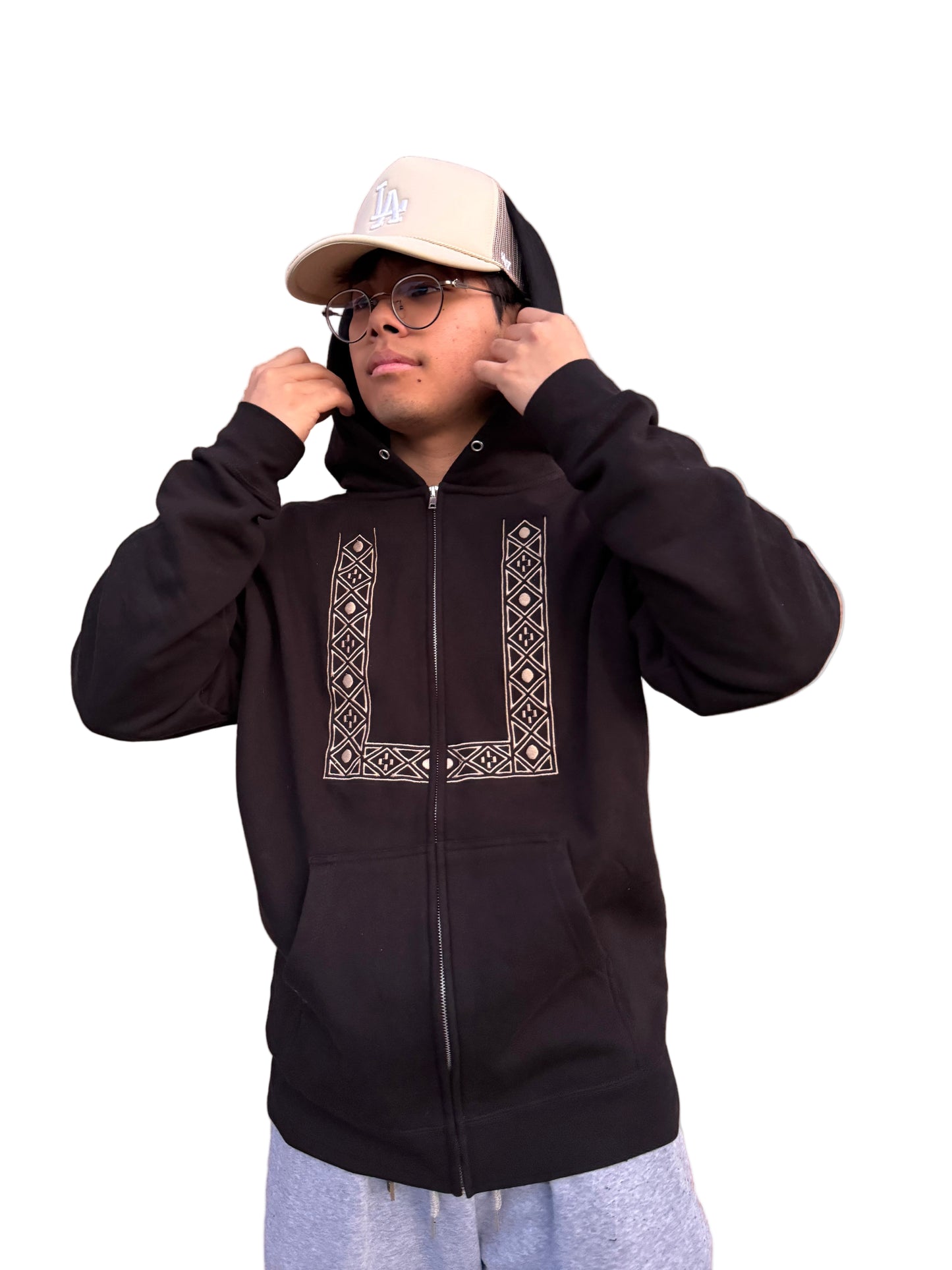 Person wearing a barong embroidered zip up sweater is adjusting his hoodie, wearing an LA baseball cap and eyeglasses