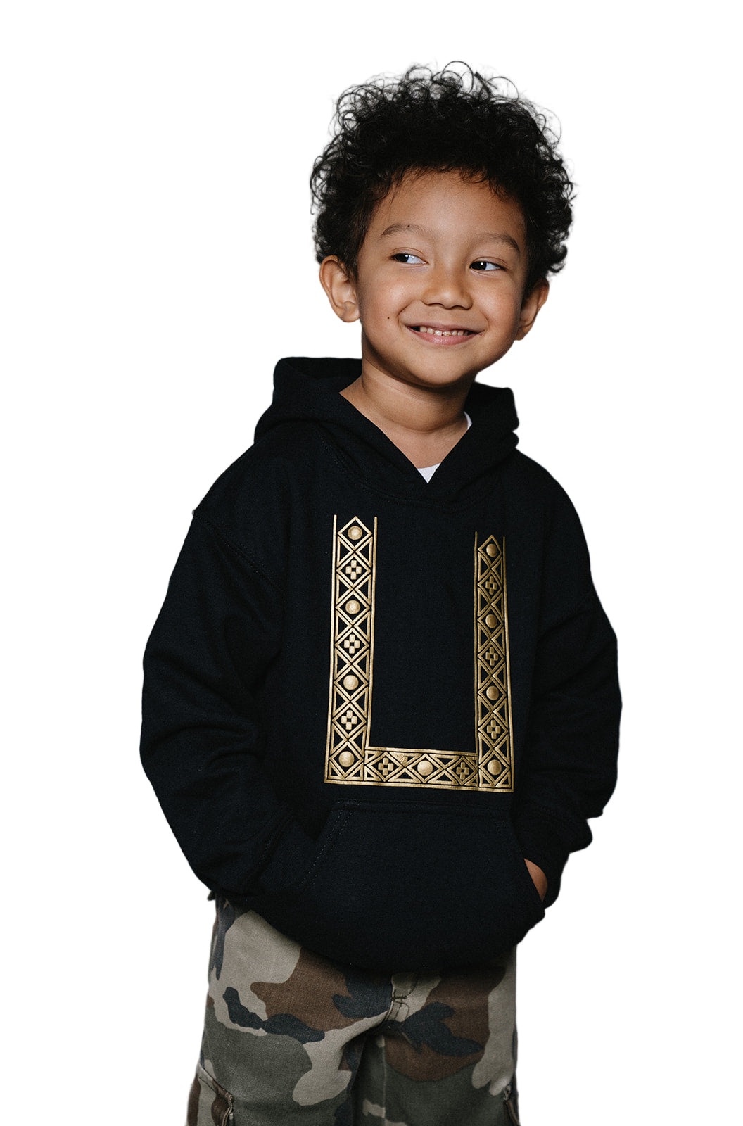Young child smiling confidently wearing a black hoodie with gold printed geometric barong pattern and camouflage cargo pants.